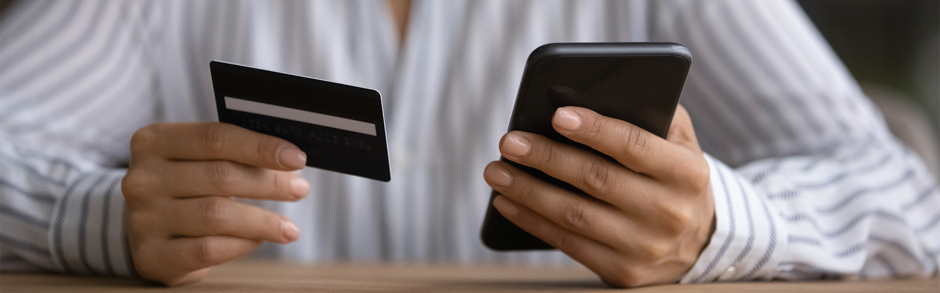 person holding a phone in their left hand and a credit card in their right, hands resting on a wooden table
